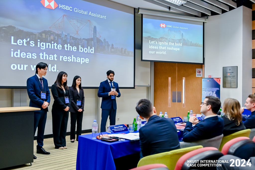 Four standing student presenters listing to feedback from four seated panelists at a presentation.